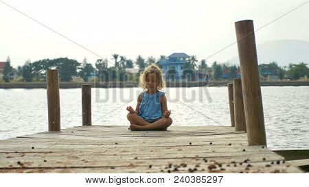 Little Child Girl Sits On The Wood Pier In Lotus Pose And Meditating