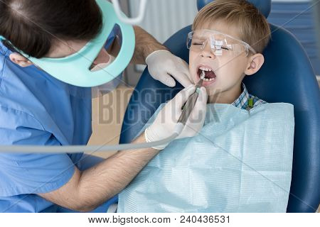 Portrait Of Cute Little Boy Sitting In Dental Chair  And Wincing With Pain While Dentist Treating Hi