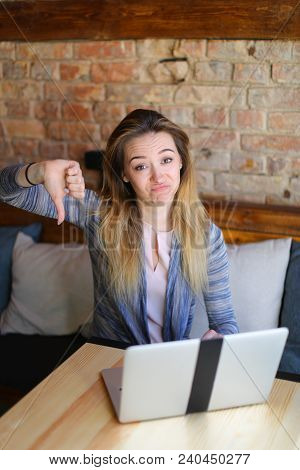 Young Woman Sitting In Cozy Coffee House Near Wooden Table With Glass Cup Of Cappuccino And Using La