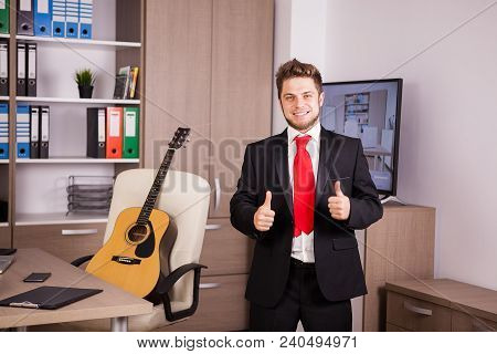 Smiling Businessman In Black Suit And Ret Tie Showing Thumbs Up To The Camera. He Is In His Office. 