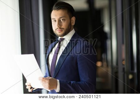 Young elegant businessman with document looking at camera while standing by office wndow