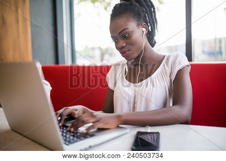 Young African American Girl In Glasses Sitting In Restaurant With Laptop. Pretty Girl With Earphones