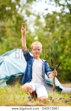 Little boyscout showing gesture of three fingers put together and looking at you in natural environment