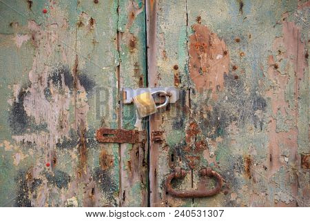 Wooden peeled door background locked with rusty handle and padlock. Timeworn brown, green entrance provides safety and privacy. Close up view with details.