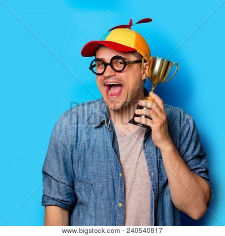 Young Nerd Man With Noob Hat Holding An Golden Cup On Blue Background