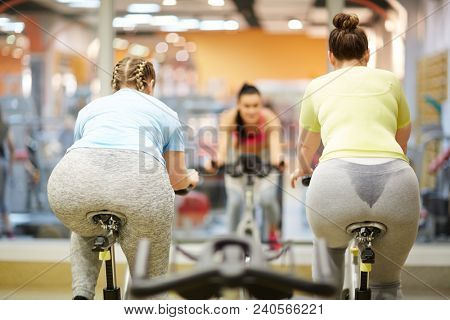 Two chubby young women in activewear cycling on bikes in front of their trainer in gym