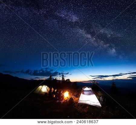Family Travellers, Four Persons Resting At Night Camping In Mountains, Sitting On Log Beside Campfir