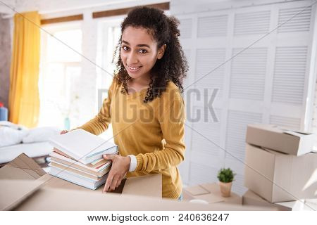 Important Books. Upbeat Young Girl Posing For The Camera And Smiling While Packing Books Into A Box 
