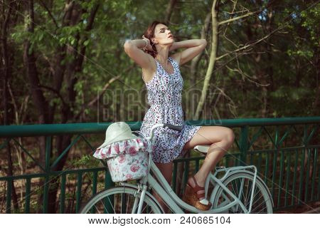 Beautiful Young Woman In A Short Dress Is Standing With A Bicycle On The Bridge.