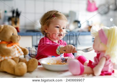 Adorable Baby Girl Eating From Fork Vegetables And Pasta. Little Child Feeding And Playing With Toy 