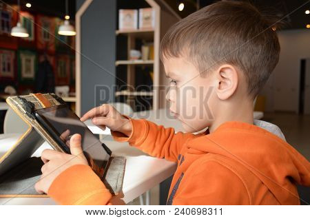 Cute Little Boy Is Sitting In A Cafe At The Table. Plays A Tablet, Watches Cartoons, Reads, Boy Play
