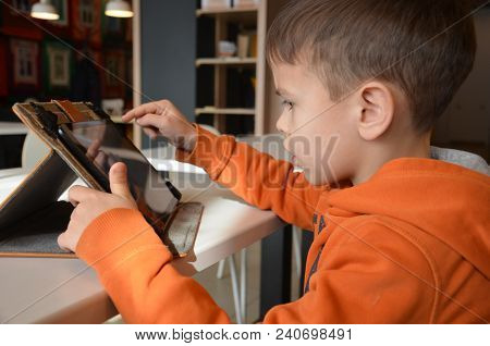 Cute Little Boy Is Sitting In A Cafe At The Table. Plays A Tablet, Watches Cartoons, Reads, Boy Play