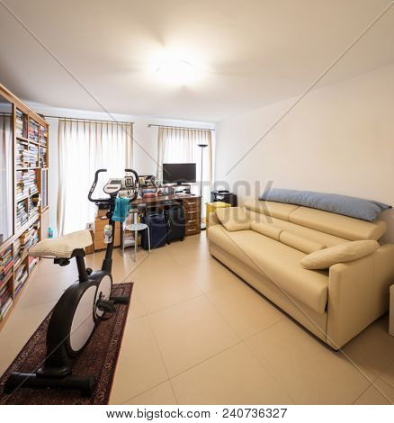 Study with an exercise bike in the middle of the room, leather sofa and bookshelf full of books. nobody inside