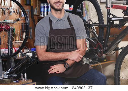 Cropped Shot Of Smiling Young Man In Aron Sitting Near Bicycles In Workshop