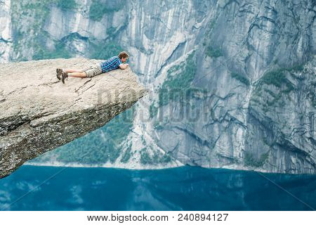 Odda, Norway - August 04, 2014: Young Man On Rock Clings To The Edge Of A Cliff And Looking Down In 