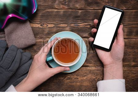 Woman Snowboarder With Blank Screen Mobile Phone Is Drinking A Hot Cup Of Tea On Mountain Resort Caf