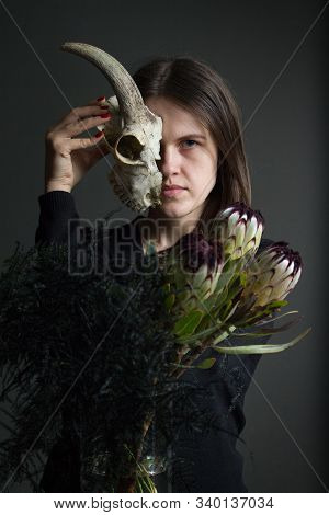 Portrait Of A Young Dark-haired Girl Holding Half A Goat Skull In Front Of Her Face And A Bouquet Of