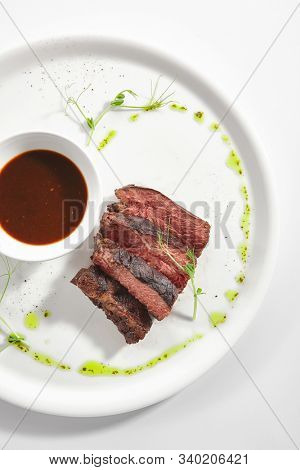 Alternative grilled skirt steak, well done machete fillet on white restaurant plate isolated. Top view of fresh juicy beef grillsteak, barbecue meat with brown spicy sauce and herbs