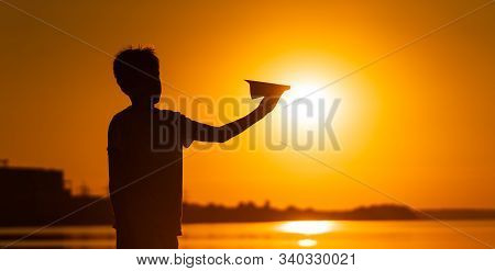 Black Silhouette Of A Boy Standing Against Orange Summer Sunset Background. Holding Small Paper Plan