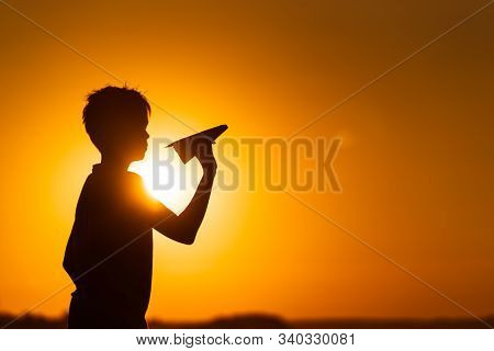 Little Boy Holds A Paper Airplane In Hand At Sunset. A Child Raised His Hand Up To Sky And Plays Wit