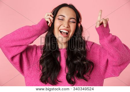 Image of optimistic beautiful asian girl with long dark hair pointing finger and listening to music with headphones isolated over pink background