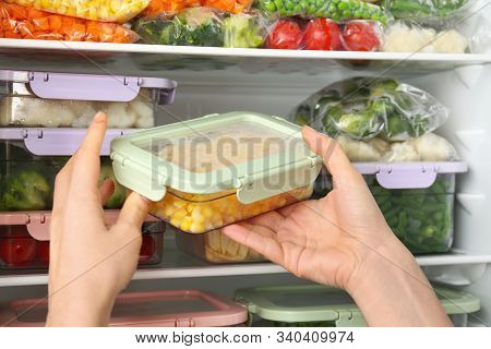 Woman Putting Container With Corn In Refrigerator With Frozen Vegetables, Closeup