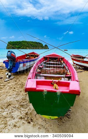 Colorful old wooden fishing boats docked by water on a beautiful beach coast. White sand sea shore landscape on tropical Caribbean island. Holiday weekend/ summer vacation setting at Pagee / Pajee Beach in Port Maria, Jamaica. Cabarita island in the dista