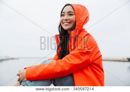 Smiling attractive young asian woman wearing raincoat spending time outdoors walking at the coastland