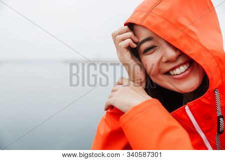 Smiling funny attractive young asian woman wearing raincoat spending time outdoors walking at the coastland, wearing a hood