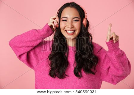 Image of optimistic beautiful asian girl with long dark hair pointing finger and listening to music with headphones isolated over pink background
