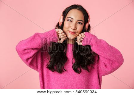 Image of optimistic beautiful asian girl with long dark hair smiling and listening to music with headphones isolated over pink background