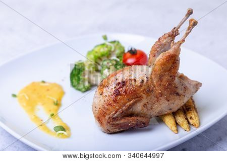 Quail Roasted Whole With Broccoli, Mini Corn, Cherry Tomato And Orange Sauce. White Plate, Light Bac
