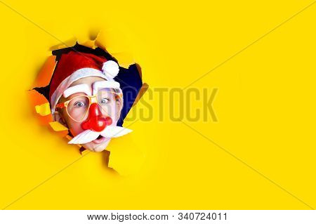Little Cheerful Santa In Glasses With A Red Nose And Mustache Getting Out Of The Ragged Yellow Backg