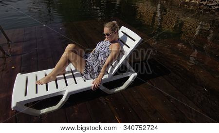 Woman Lie On A Sunbed In Sunglasses And A Boho Silk Shawl. Girl Rest On A Flood Wood Underwater Pier