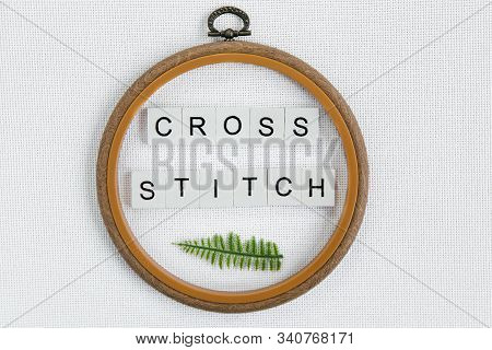 Minimalistic Flat Lay: A Wooden Hoop, The Inscription Cross Stitch And A Sprig Of Fern On A White Ca