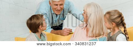 Panoramic Shot Of Senior Man Talking To Happy Wife Sitting On Sofa With Grandchildren