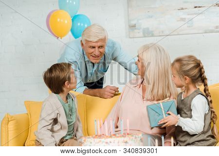 Smiling Senior Man Talking To Happy Wife Sitting On Sofa With Grandchildren