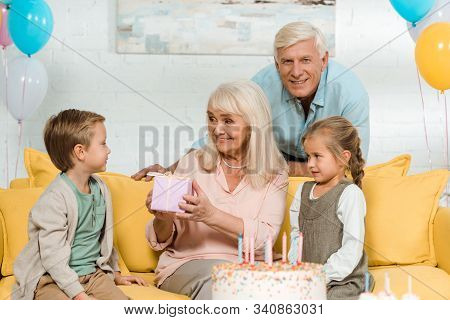 Happy Senior Woman Holding Gift Box While Sitting On Yellow Sofa Near Grandchildren And Husband