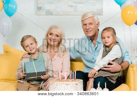 Happy Grandparents Sitting On Sofa With Grandchildren And Smiling At Camera
