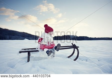 Beautiful Winter. Little Baby Girl With Red Cap Sit On Old Wooden Sledge In Snow-covered Landscape