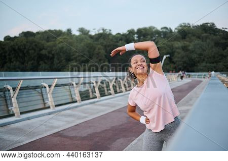 Happy Sporty Woman Smiles Toothy Smile, Exercising Outdoors On The Bridge, Focusing On Stretching Du