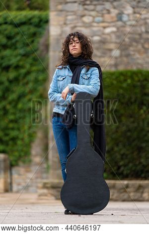 Young Woman With Curly Hair And Glasses Posing With A Guitar Case In The Street