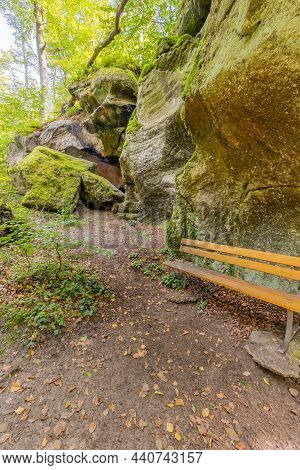 Dirt Path, Wooden Bench Next To Huge Sandstone Rock Formation With Moss, Green Trees In The Backgrou