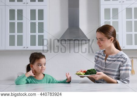 Mother Feeding Her Daughter In Kitchen. Little Girl Refusing To Eat Vegetables