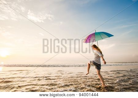 Cheerful young girl with rainbow umbrella having fun on the beach before sunset. Travel, holidays, v