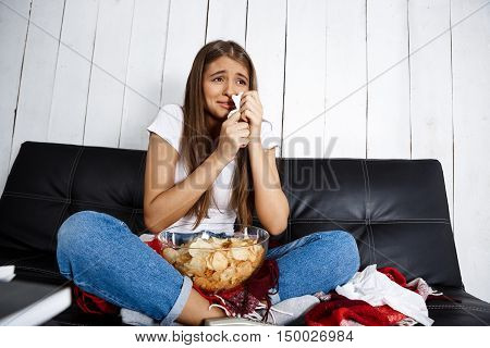 Sad young beautiful girl watching melodrama, crying, sitting on sofa at home. Copy space.