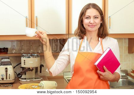 Relax and rest in kitchen. Middle aged woman chef housekeeper holding cookbook with cup of tea coffee. Housewife resting at home.