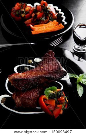 fresh red beef meat steak barbecue garnished vegetable salad sweet potato and basil on black plate over black wooden table with bbq sauce in sauceboat