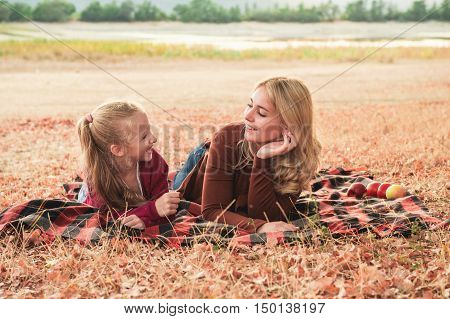 Close up portrait of two smiling sisters lying under tree on a plaid. Young mom and cute daughter have a good time outsaide. Autumn weather falling leaves.