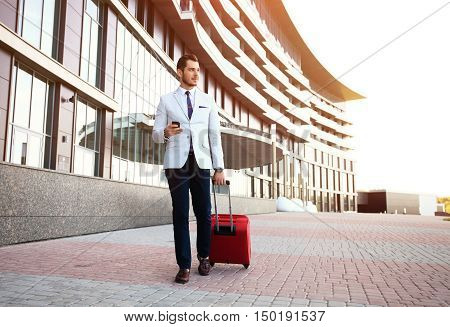 Young happy traveler businessman making call after arriving at hotel outside with his luggage. Smiling traveler using smartphone